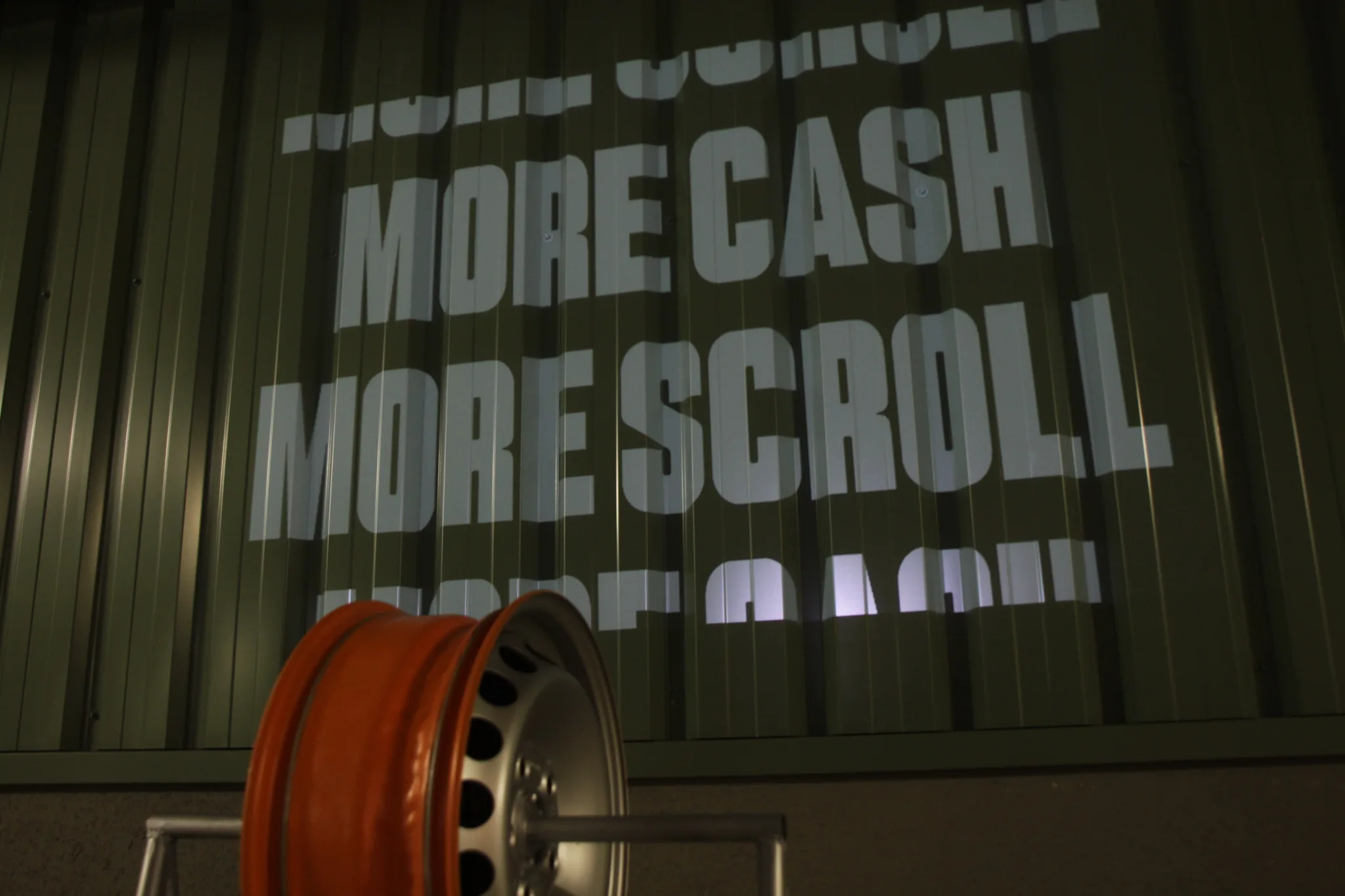 Orange wheel rim with MORE CASH MORE SCROLL projected in white on green corrugated metal wall