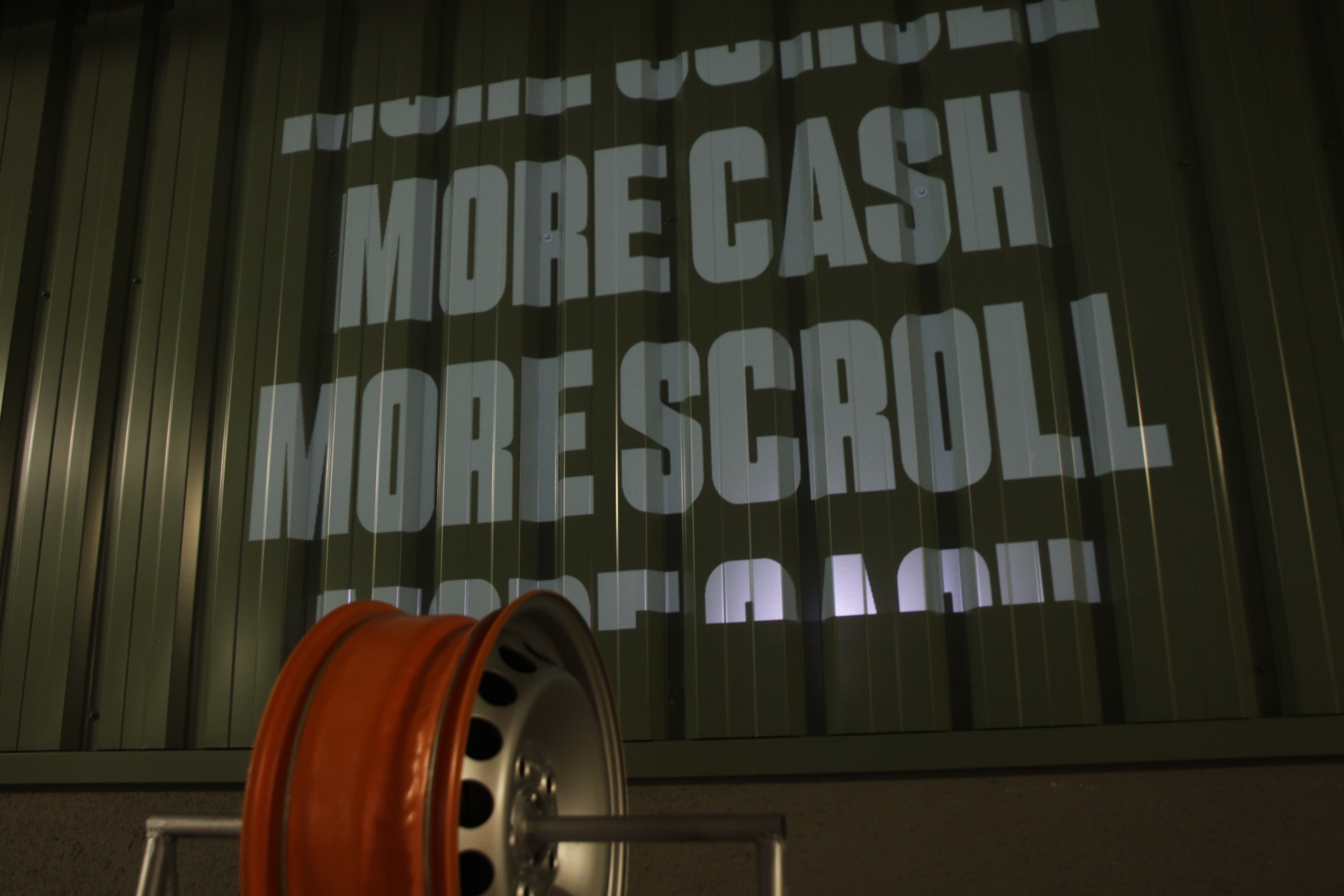 Orange wheel rim with MORE CASH MORE SCROLL projected in white on green corrugated metal wall