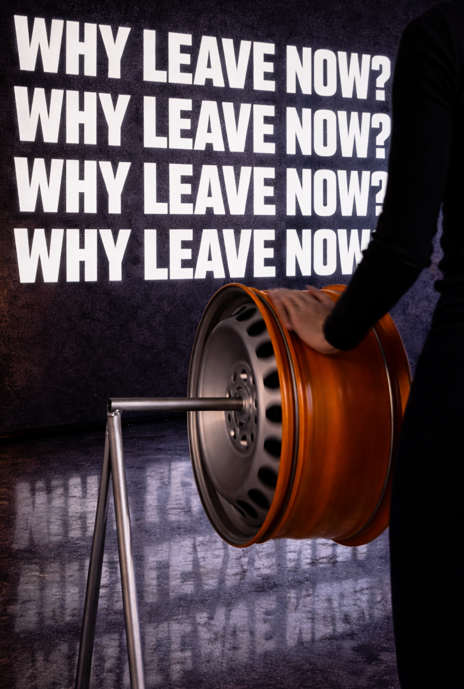 Person spinning orange wheel with WHY LEAVE NOW? projected in bold white text on dark wall